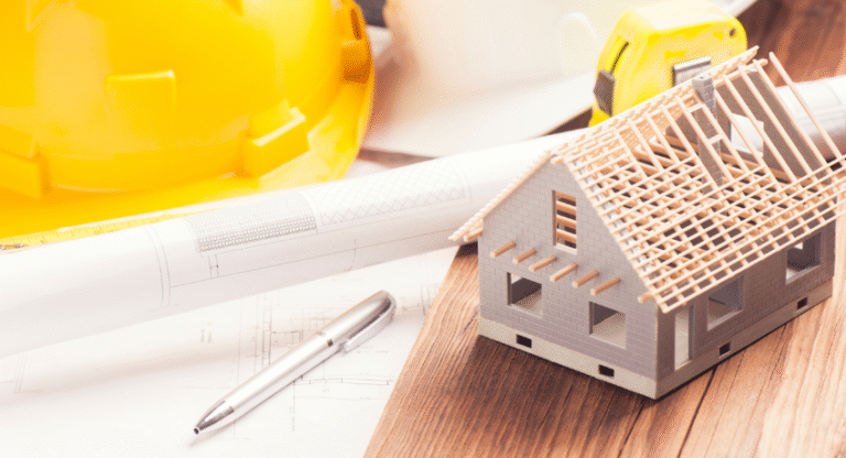 Maquete de casa com estrutura do telhado sobre mesa com planta arquitetônica, capacete de obra e ferramentas.