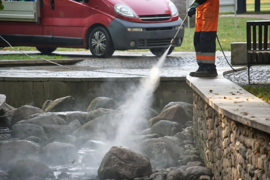 Hidrojato de alta pressão: Como funciona e quais são as aplicações na limpeza industrial: Operador de limpeza com jato de água de alta pressão usando mangueira sobre pedras em uma canaleta externa, com veículo ao fundo. A limpeza remove sujeira e detritos com vapor e água sob pressão.